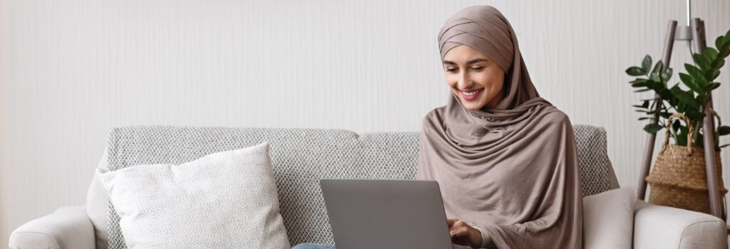 Modern arab girl in hijab using laptop at home, working remotely while sitting on couch in living room